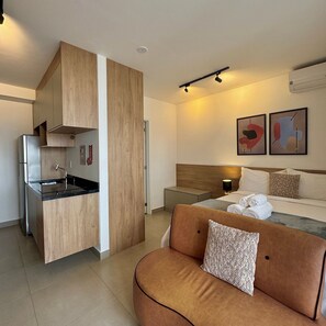 A compact hotel room with a kitchenette, a bed with a headboard, a bedside table, and two abstract wall art pieces.