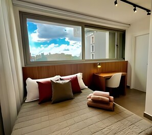 Une chambre moderne dotée d’une grande fenêtre, d’un bureau et d’un coin salon confortable avec des coussins.