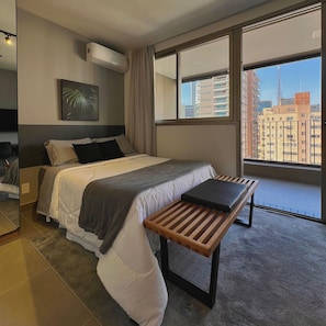 A modern hotel room with a large bed, a bedside table, a wall-mounted air conditioner, and a window with a city view. - Apartamentos Prime-Prox a Oscar Freire (São Paulo)
