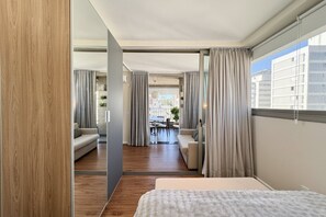 Egy modern szállodai szoba, melyben egy nagy ágy, egy kanapéval és karosszékekkel berendezett ülősarok, egy étkezőasztal található, és a nagy ablakokon keresztül kilátás nyílik a városra.