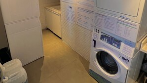 A laundry room with a front-loading washing machine, a dryer, and a utility cart with a detergent container. Instructions and symbols are displayed on the wall. - Apartamentos Prime-Prox a Oscar Freire (São Paulo)