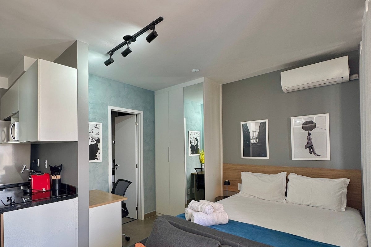 Ett modernt hotellrum med ett litet kök, en säng med vita sängkläder, en grå soffa med orange kuddar och konstverk på väggarna.