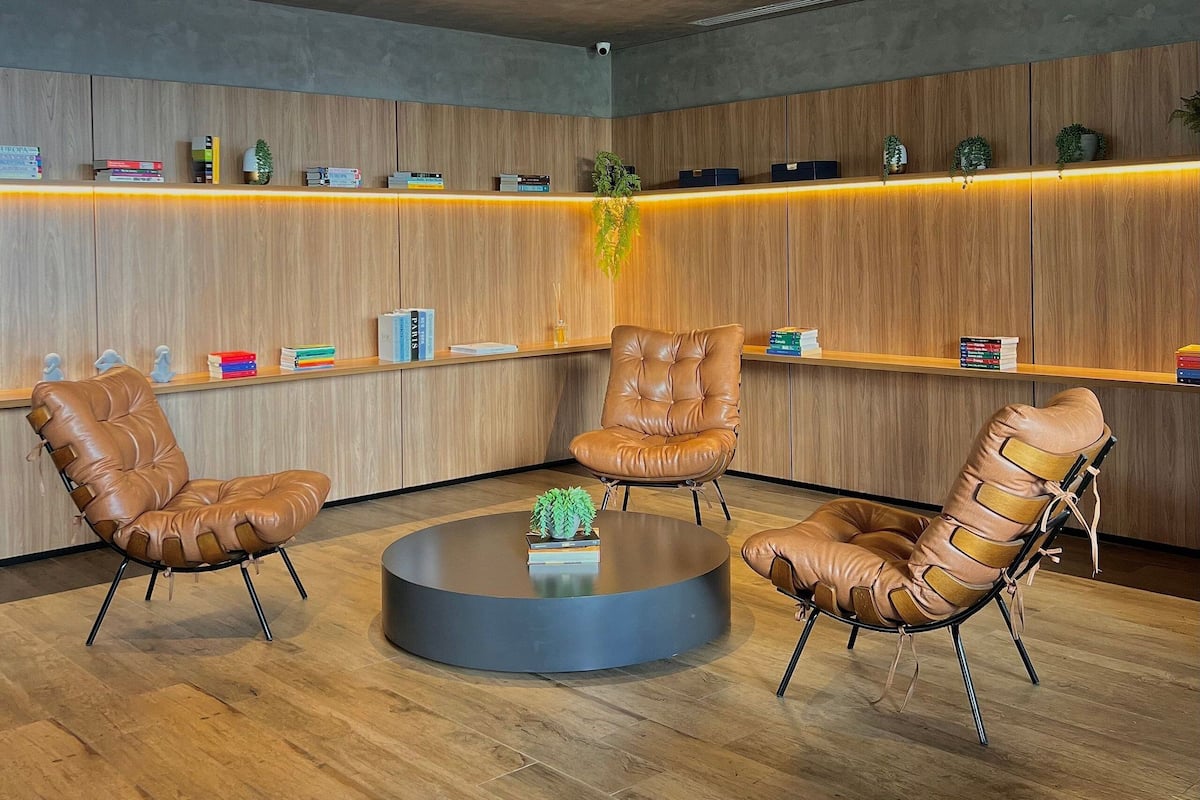 Uma área de espera moderna com cadeiras de couro, uma mesa redonda e prateleiras de madeira com livros e objetos decorativos.