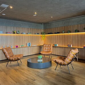 Uma área de espera moderna com cadeiras de couro, uma mesa redonda e prateleiras de madeira com livros e objetos decorativos.