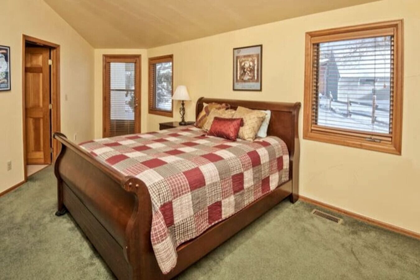 A bedroom with a wooden bed, a checkered bedspread, a nightstand, a lamp, and a window with blinds.