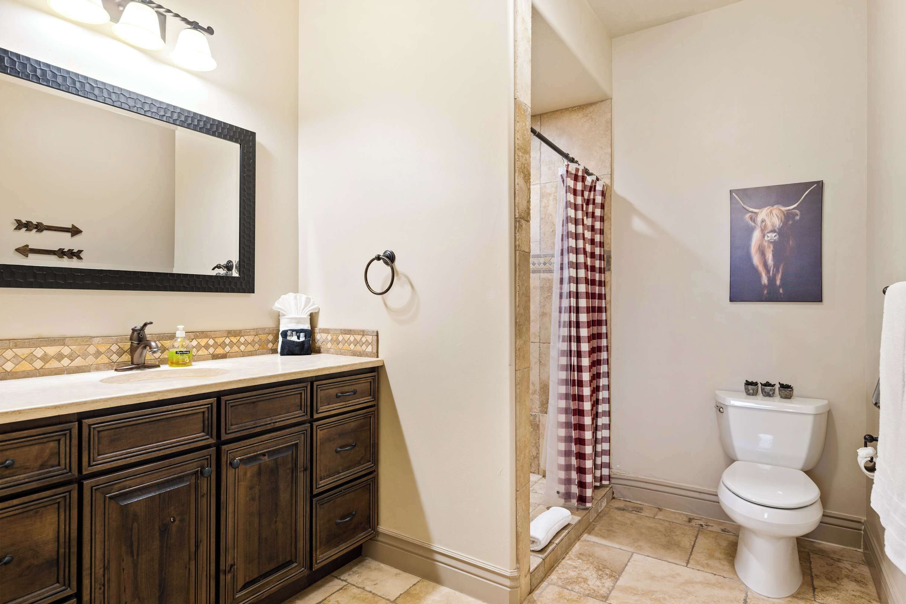 A bathroom with a dark wood vanity, a white toilet, a shower with a checkered curtain, and a mirror with lights.