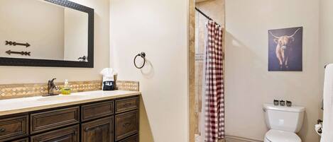A bathroom with a dark wood vanity, a white toilet, a shower with a checkered curtain, and a mirror with lights.