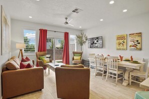 Sala de estar com sofá, poltronas, mesa de jantar com cadeiras, ventilador de teto e janela com cortinas.