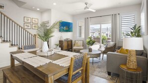 Ein geräumiger Wohnbereich mit Esstisch, Stühlen, einer Couch und einem Couchtisch. Eine Treppe mit Geländer, ein Deckenventilator und ein großes Fenster mit Jalousien sind ebenfalls vorhanden.