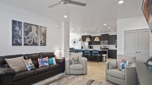 Ein modernes Wohnzimmer mit einem dunkelbraunen Ledersofa, zwei grauen Sesseln und einem Streifen Teppich. Im Hintergrund ist ein Essbereich mit Bar und Küche erkennbar.