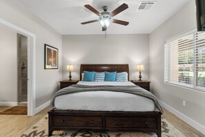 A bedroom with a wooden bed, two bedside tables with lamps, a ceiling fan, a window with blinds, and a door leading to a bathroom.
