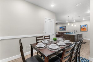 A dining area with a wooden table set for four, surrounded by chairs. There is a kitchen area in the background with a brown countertop and a sink.