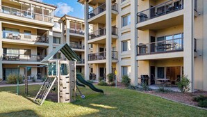 Un immeuble résidentiel à plusieurs étages doté de balcons, d’une petite aire de jeux avec un toboggan et d’espaces extérieurs aménagés pour s’asseoir.