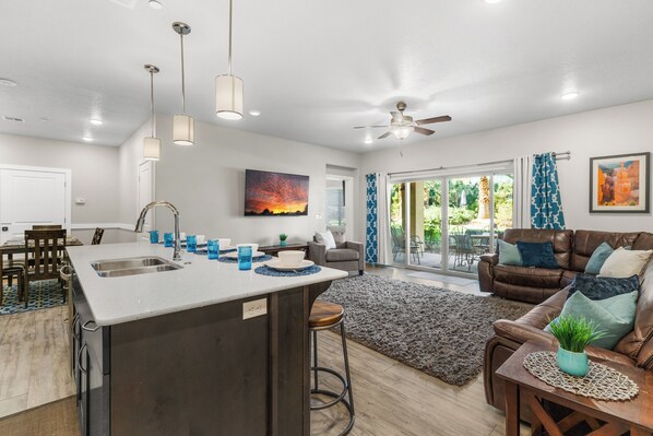 A modern kitchen with a central island, a dining area with a table and chairs, a living room with a sofa and a coffee table, and a large window with curtains.