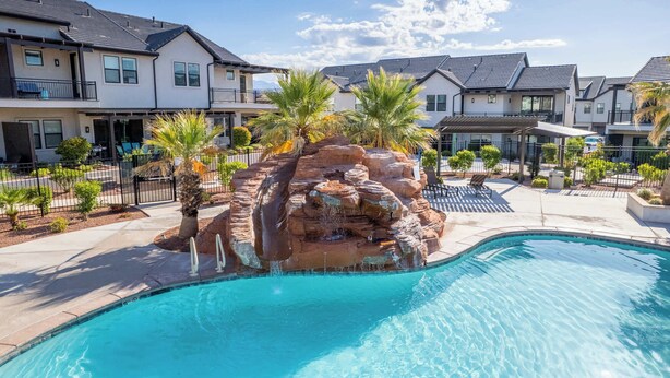 A swimming pool with a rock waterfall feature, surrounded by modern houses with balconies and palm trees.