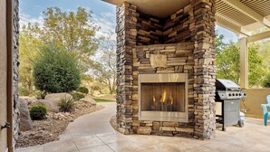 A stone fireplace with a fire, a grill, and a patio area with a tiled floor and landscaping. - Sweet Retreat | Rest & Relax Near National Parks (Hurricane)