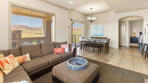 A spacious living room with a brown sofa, a dining area with a table and chairs, and a view of the outdoors through large windows. - Desert Ocean Vibes- Resort Amenities (Hurricane)