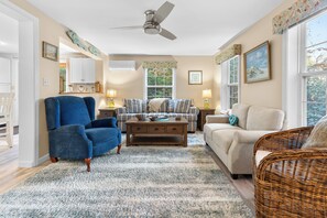 A living room with a blue armchair, a sofa, a coffee table, and a wicker chair. There is a ceiling fan, a window with a view of trees, and a painting on the wall.