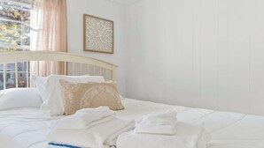 A neatly made bed with pillows and folded towels, a decorative wall piece, and a window with curtains. - Cappawack Cottage | Firepit & Private Beach Nearby (Mashpee)