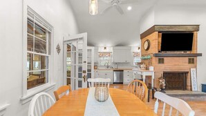 A spacious kitchen with white cabinets, a wooden dining table, and a built-in TV. - Cappawack Cottage | Firepit & Private Beach Nearby (Mashpee)