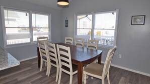 A dining area with a wooden table and six chairs, large windows with blinds, and a hanging pendant light.