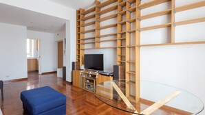 A modern living room with a glass coffee table, wooden shelving, a flat-screen TV, and a blue ottoman. - Vittorio Panoramic Apartment (Roma)