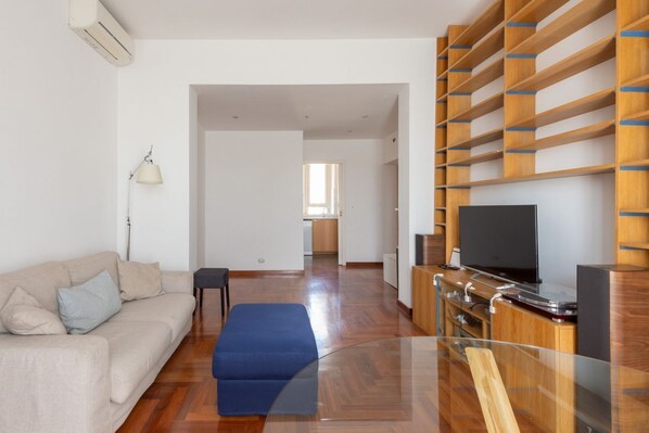 A modern living room with a sofa, ottoman, TV, and wooden shelving unit.