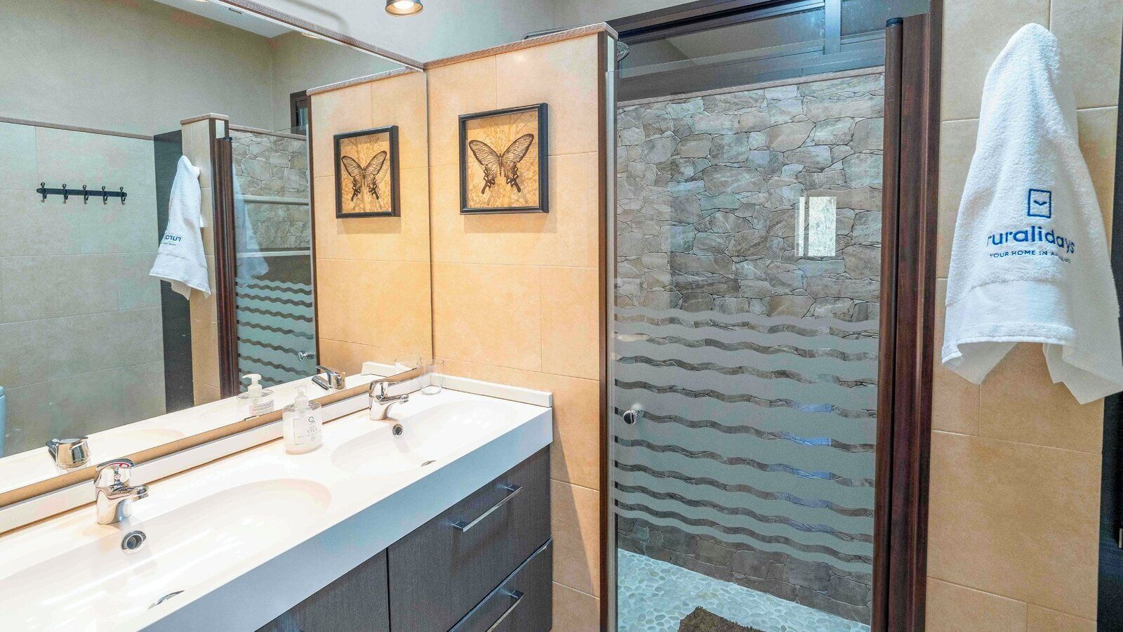 Un baño moderno con lavamanos doble, cabina de ducha de vidrio y cuadros con mariposas en la pared.