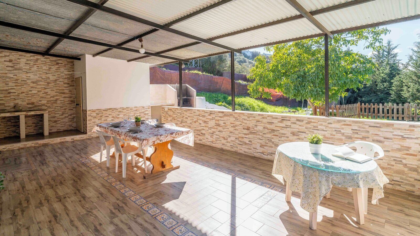 En udendørs terrasse med stenvæg, et træbord med stole og et glastop med en stol.