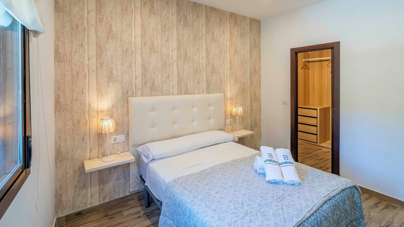 Un dormitorio con pared de paneles de madera, cabecera capitonada, mesita de noche con lámpara y cama con colcha estampada y toallas enrolladas.