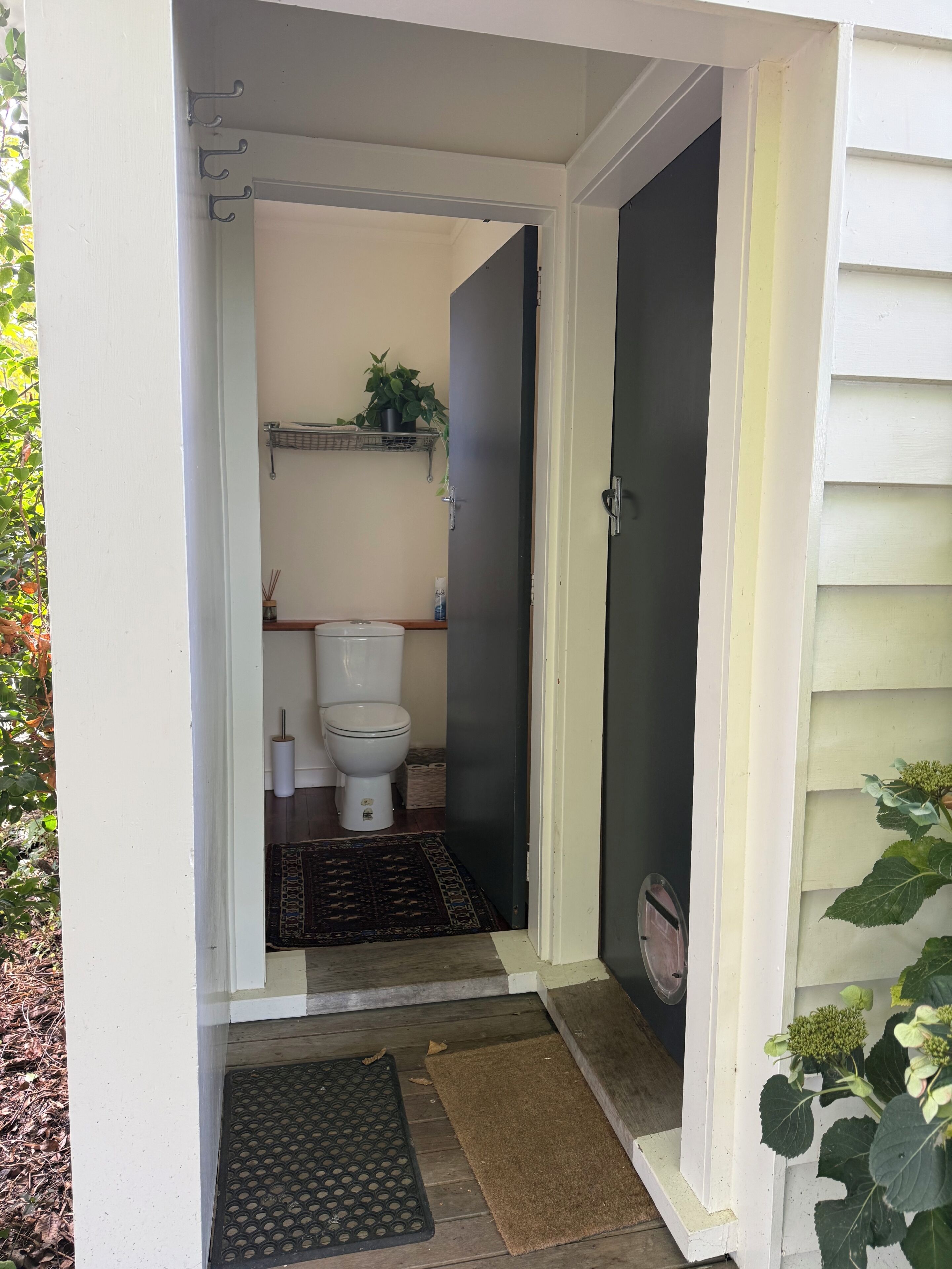 A small, enclosed bathroom with a toilet, a shelf with a plant, and a door leading to a porch.