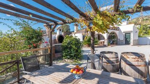 A wooden deck with a table holding a fruit bowl, surrounded by rustic barrels and a pergola with vines.
