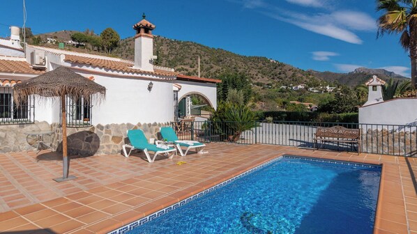 A swimming pool with two lounge chairs and a table, surrounded by a stone wall and a fence.