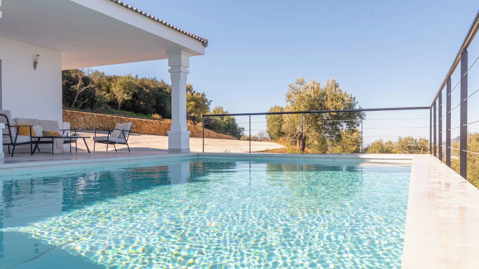 A swimming pool with a covered area and lounge chairs, surrounded by a stone wall and trees.