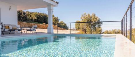 A swimming pool with a covered area and lounge chairs, surrounded by a stone wall and trees.