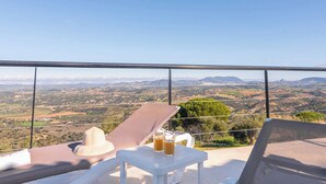 Een balkon met een helder uitzicht op een bergachtig landschap, een witte tafel met een drankje en een zonnehoed op de stoel.