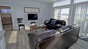 Ein Wohnzimmer mit einer schwarzen Ledersofa, einem Fernseher und einem Couchtisch. Ein Fenster bietet Blick auf den Strand.