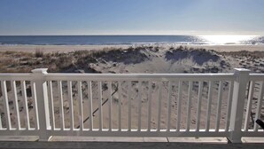 Blick auf einen Strand von einer Holzterrasse mit weißer Reling, Sanddünen und dem Meer im Hintergrund.