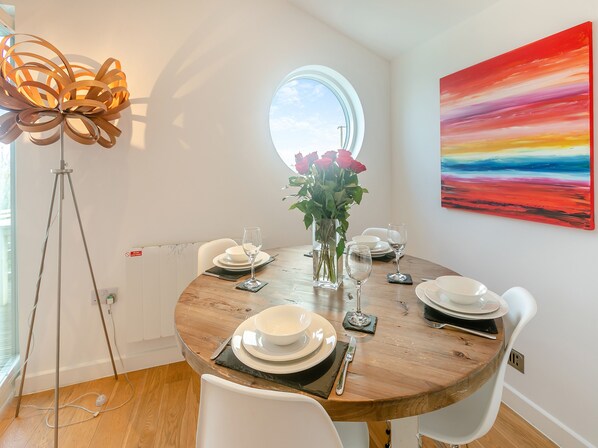 A dining area with a round table, white chairs, a vase with flowers, and a painting on the wall.