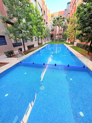 A swimming pool with a tiled floor, surrounded by greenery and buildings. - Appartement Cosy Vus piscine Familiale (Marrakech)