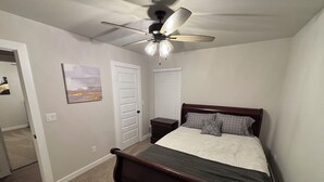 Une chambre à coucher comprenant un lit, un ventilateur de plafond, une table de chevet, une porte et un tableau accroché au mur.