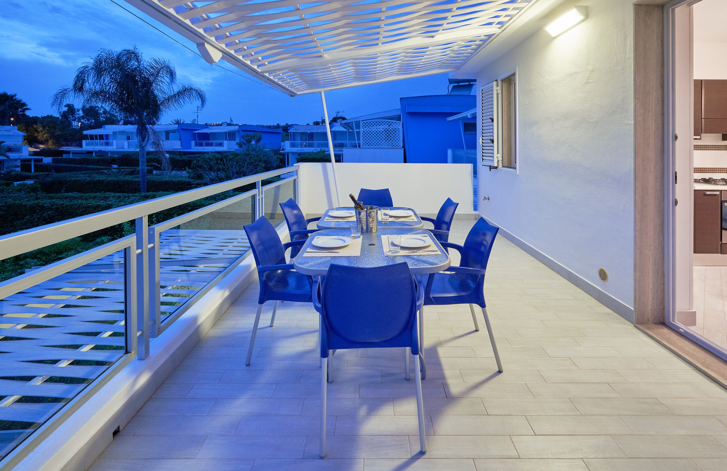 Eine Terrasse mit Tisch für zwei Personen, umgeben von blauen Stühlen, mit Blick auf einen Garten und Nachbargebäude.