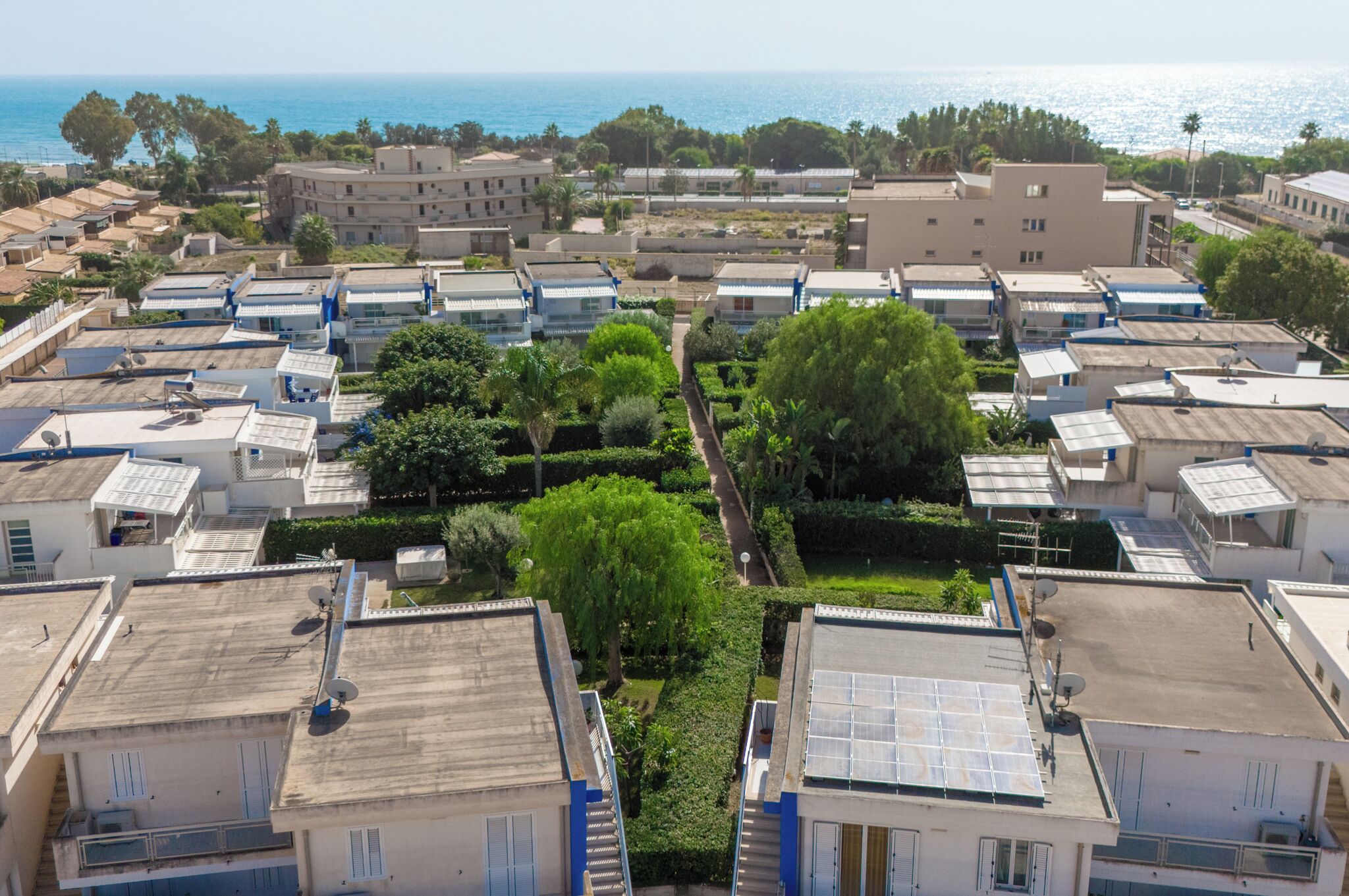 A residential area with multiple buildings, some with solar panels, and a clear view of the sea in the background.
