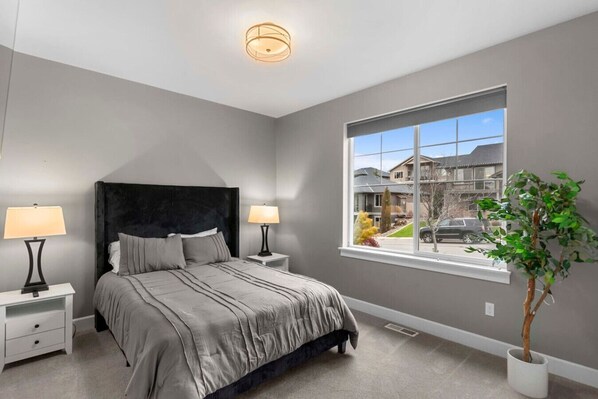 A bedroom with a large bed, two bedside tables with lamps, a window with a view of buildings, and a potted plant. - Luxury Escape in East Wenatchee (East Wenatchee)