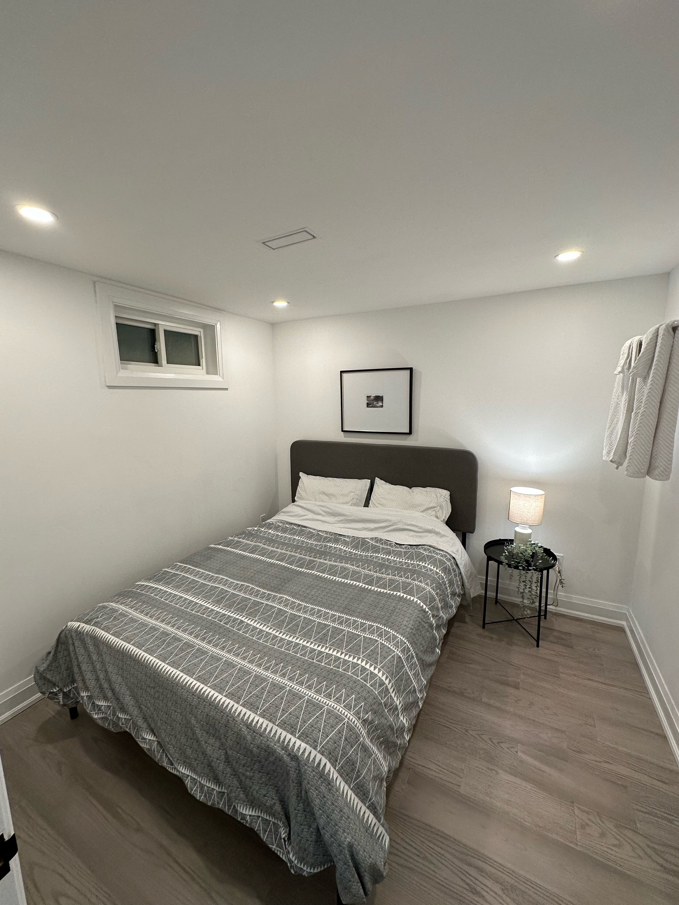 Une chambre à coucher avec un lit, une petite table de chevet avec une lampe, un tableau encadré au mur et une fenêtre.