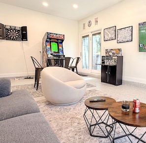 A living room with a dartboard, a dart machine, a white armchair, a sofa, a coffee table, and a cabinet.