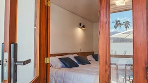 A bedroom with a bed, a nightstand, a wall-mounted light, and a window with a view of palm trees. - Pousada Mirante do Sol (Paraty)