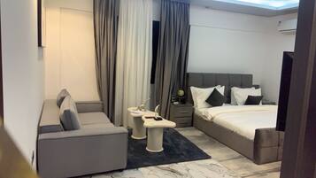 Ein modernes Hotelzimmer mit grauem Sofa, einem Bett mit weißer Bettwäsche, einem kleinen Nachttisch und einem großen Fenster mit Vorhängen.