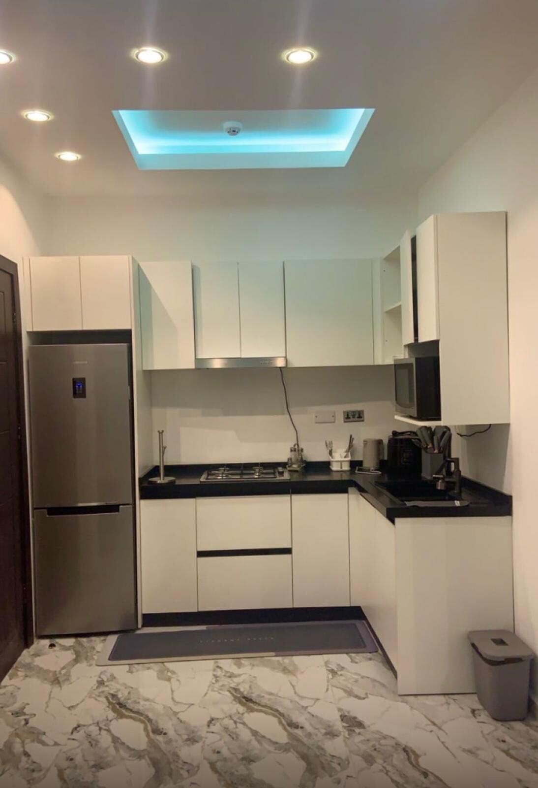 A modern kitchen with white cabinets, a stainless steel refrigerator, a black countertop, and a marble floor.