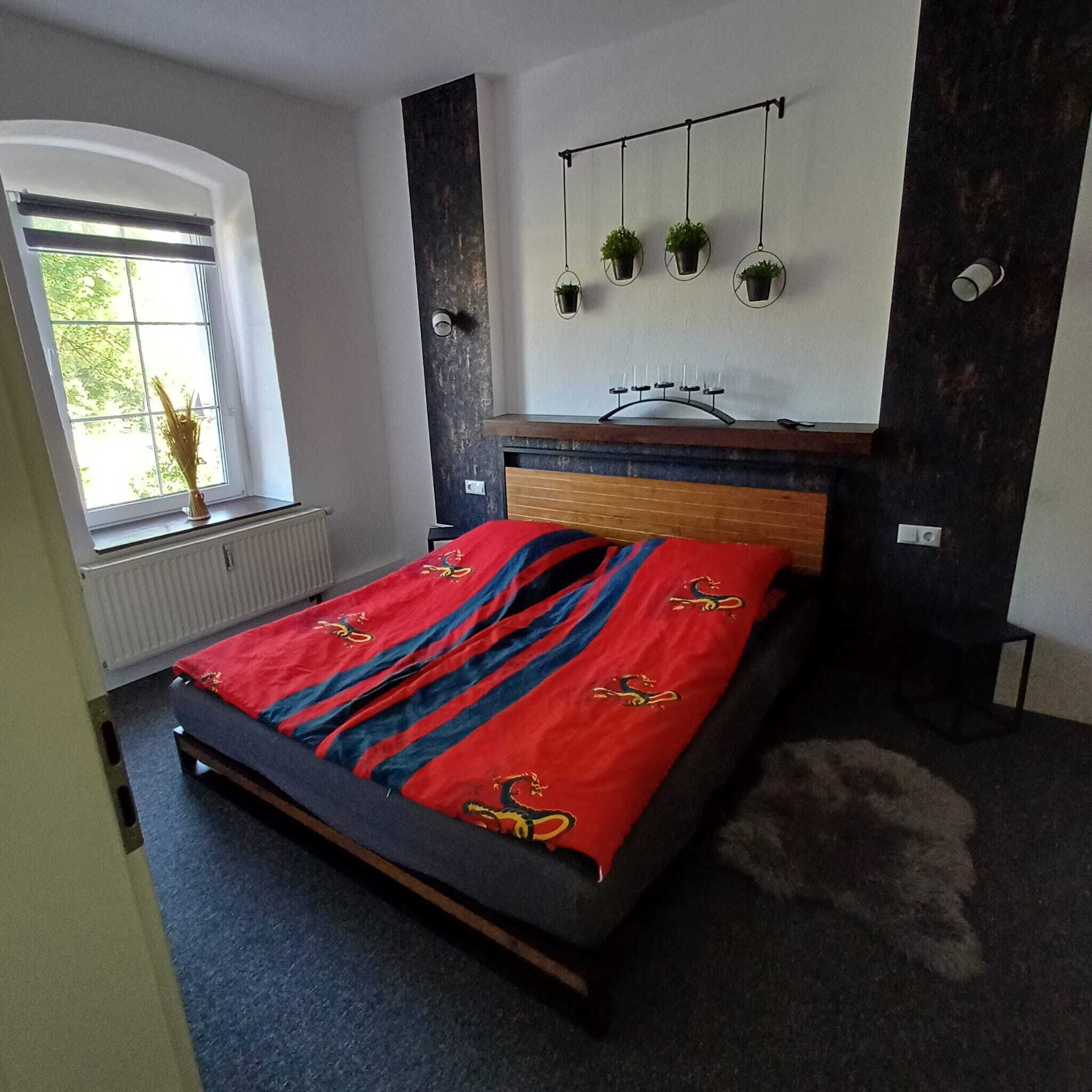 A modern bedroom with a bed, a window, a radiator, a wall-mounted shelf with plants, and a dark accent wall.